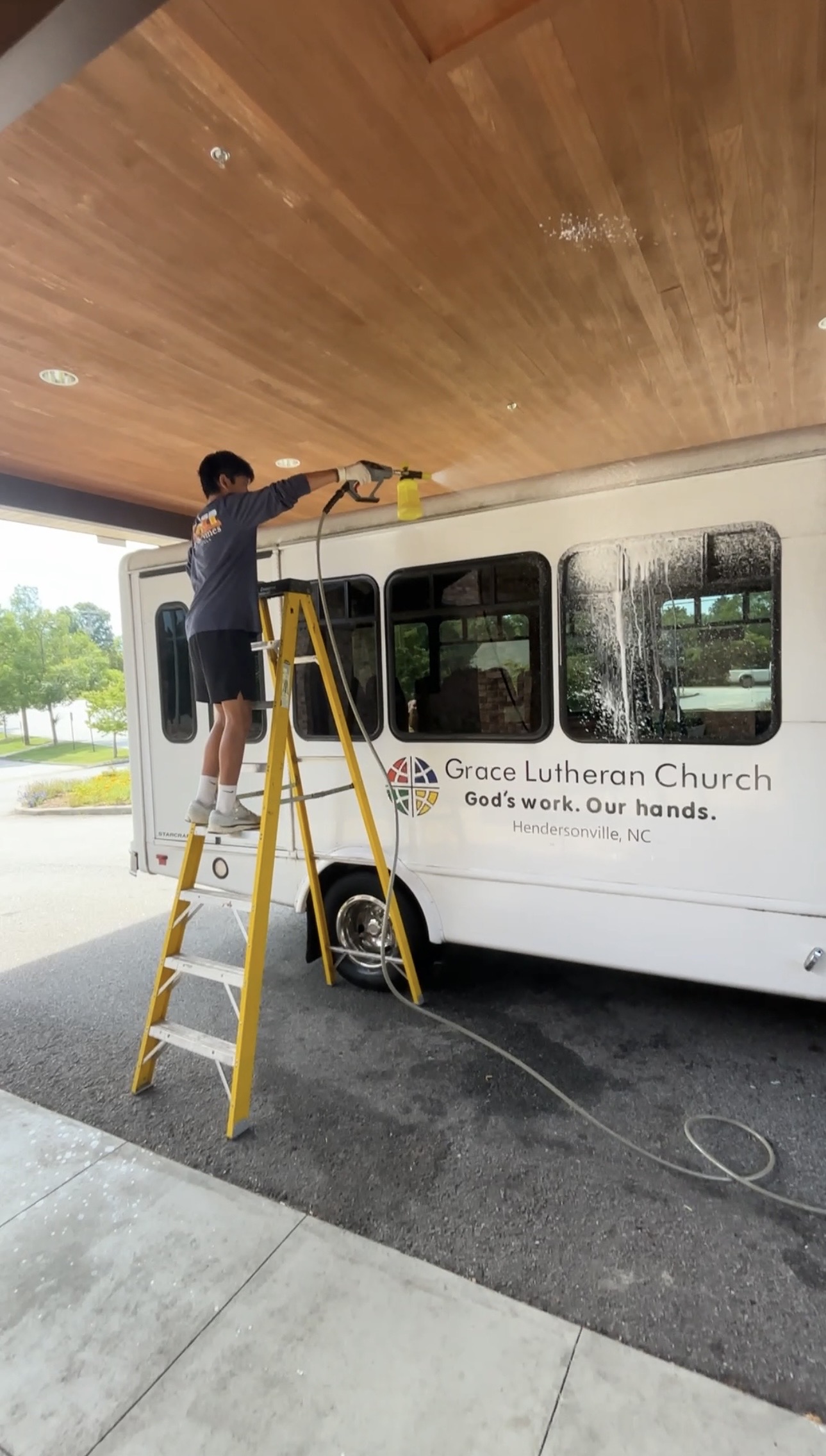 Detailer foaming the upper windows of a church shuttle bus from a ladder
